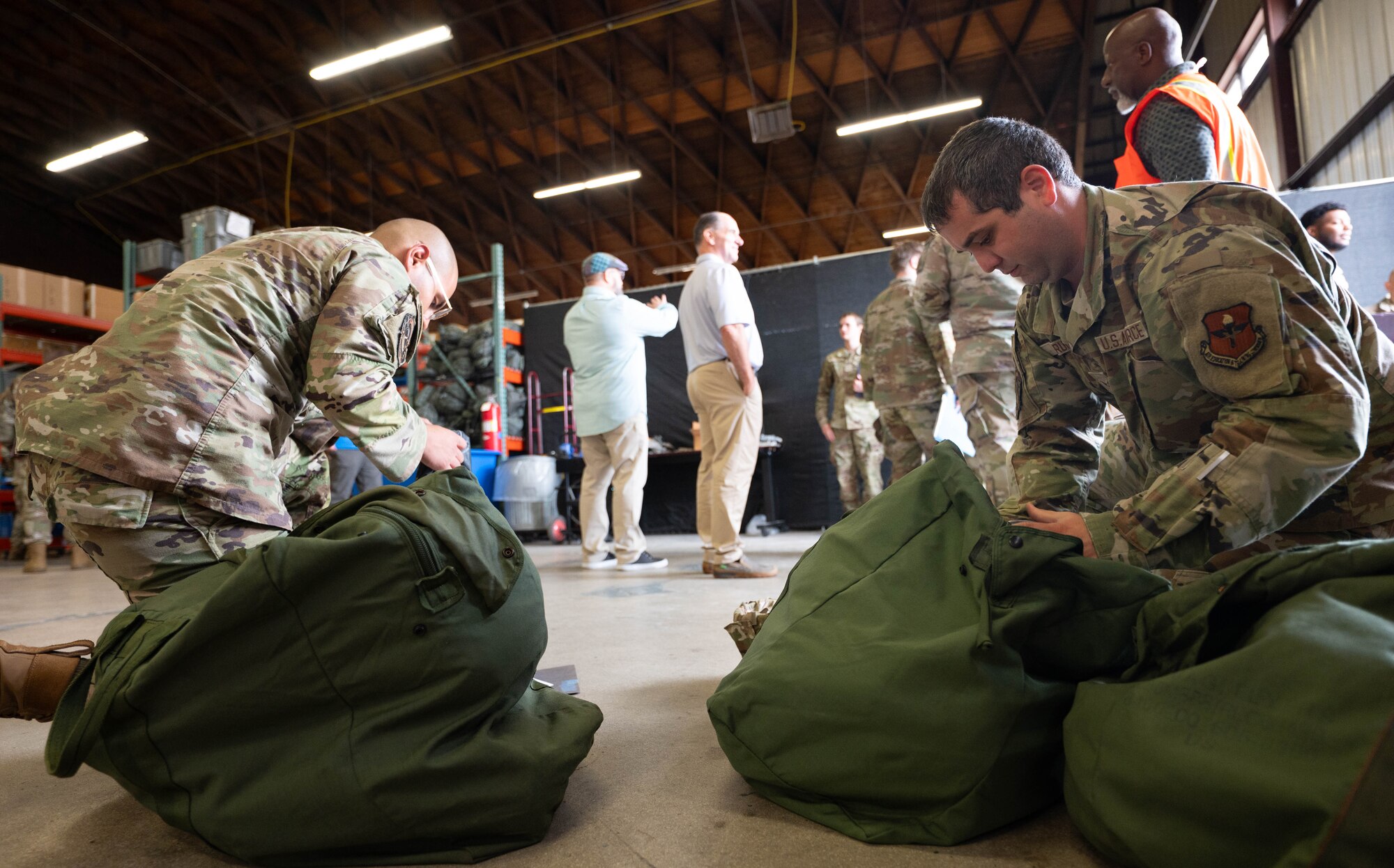 Two men in Air Force camouflage uniforms kneel down by their green duffle bags to inspect their gear.