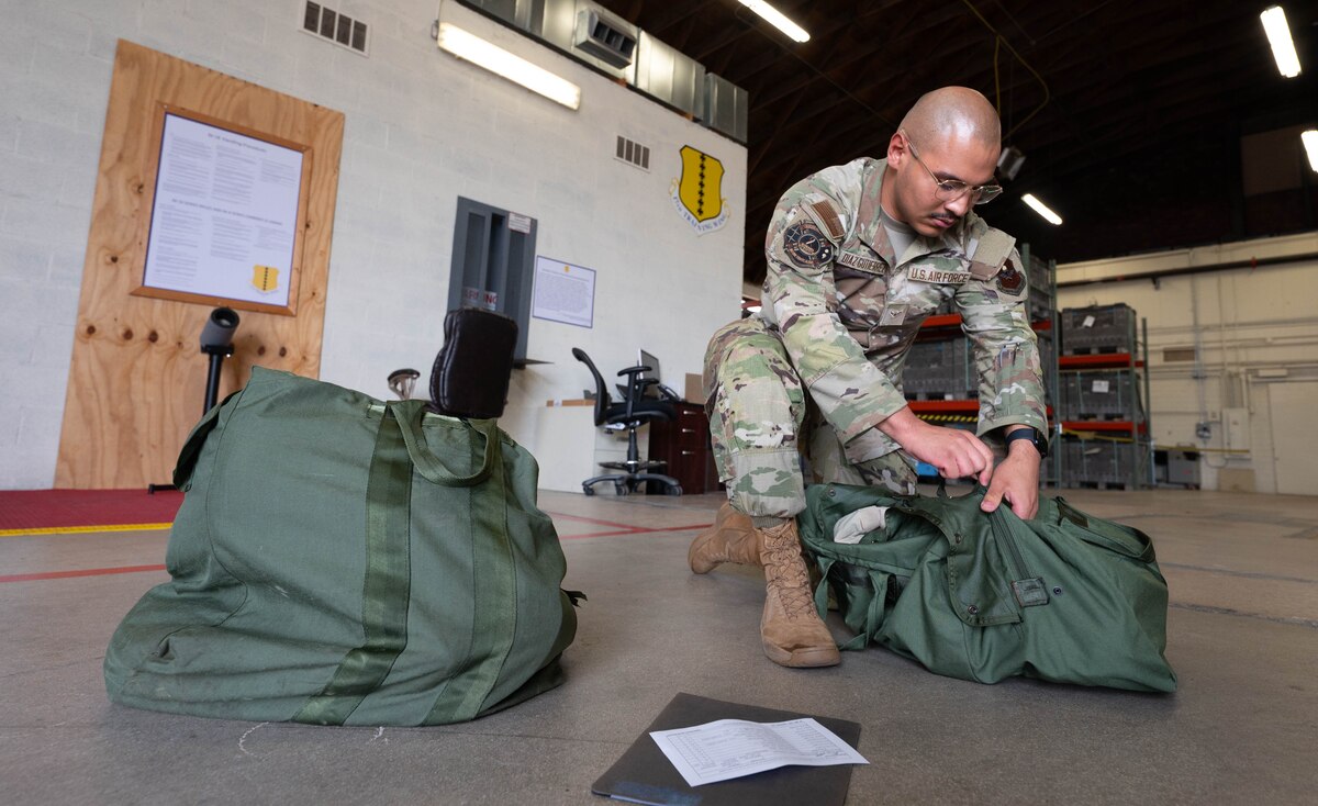 A man in an U.S. Air Force occupational camouflage uniform kneels down to zip up a green duffle bag during an exercise.
