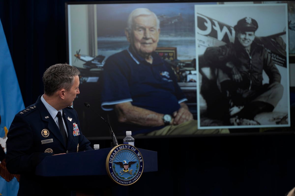 An airman in dress uniform stands behind a podium and looks toward his left at a projector screen with two images of a retired service member, one image of an elderly white man with white hair and the other a photo of the same man in a bomber jacket in front of a plane when he was younger.