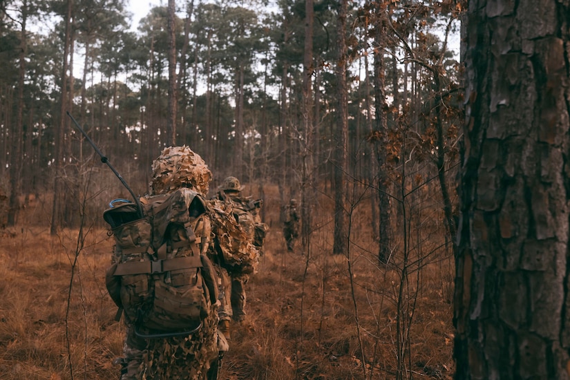 Three people wearing camouflage military uniforms and tactical gear walk through the woods.