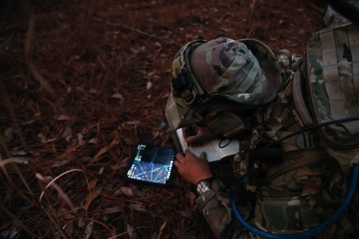A person wearing a camouflage military uniform lies on the ground in the woods while writing coordinates from a GPS into a notebook.