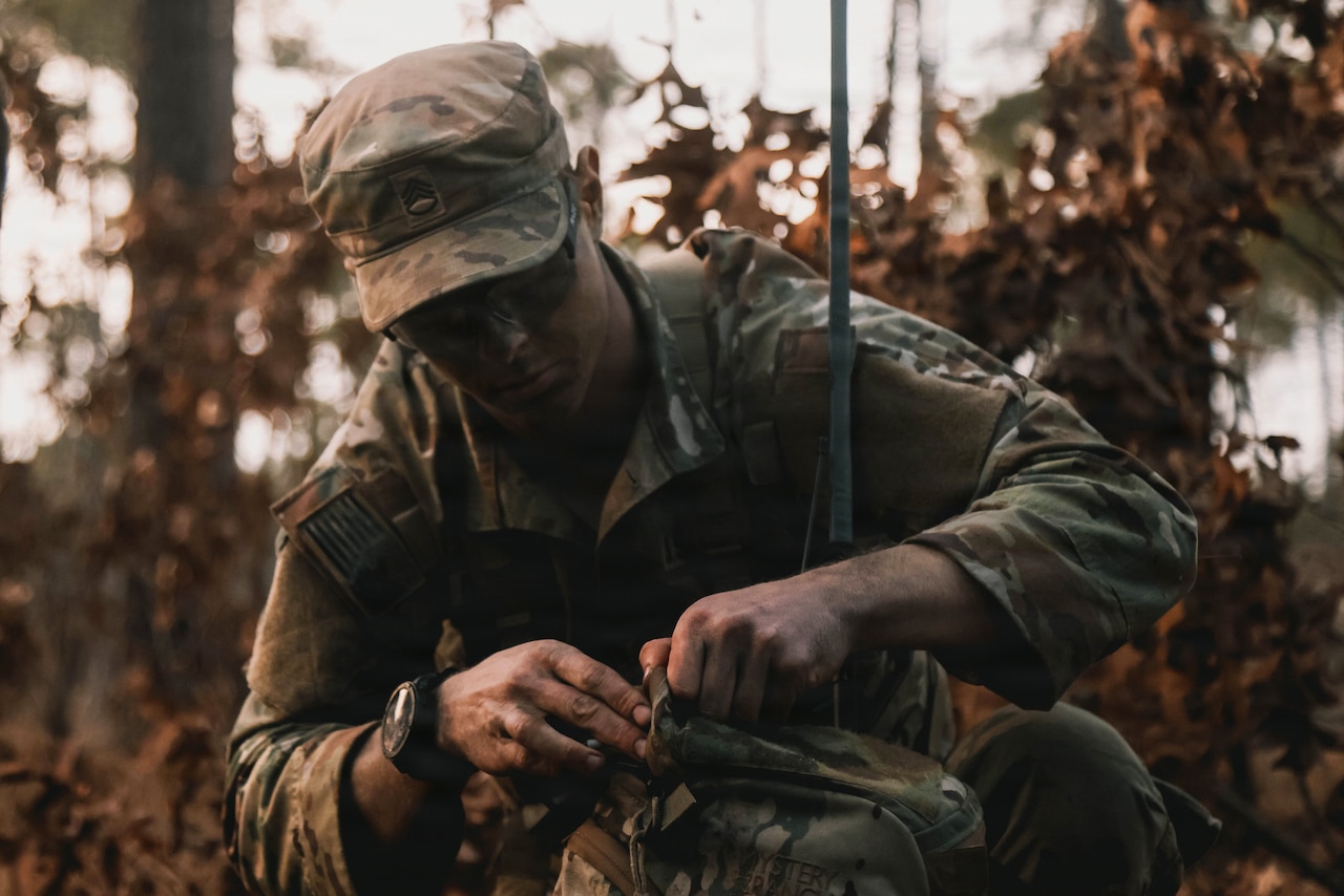 A man wearing a camouflage military uniform kneels in the woods while checking his equipment.