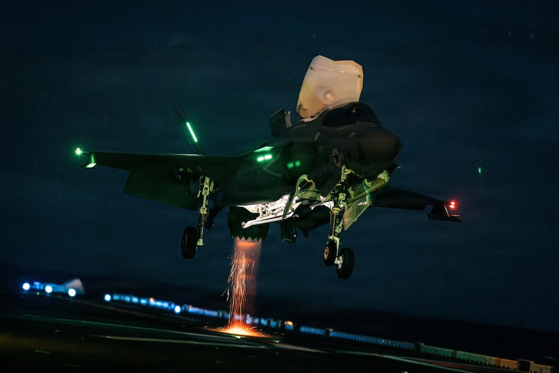 An aircraft prepares to land on a ship in the dark illuminated by green, orange and red lights.