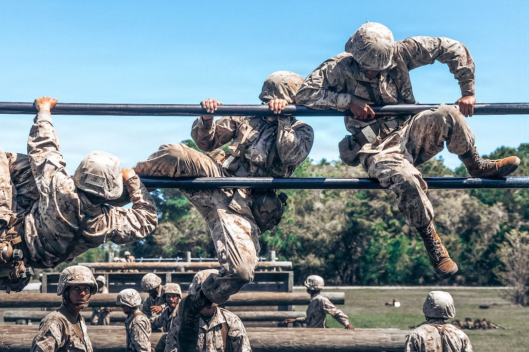 Three Marine Corps recruits at different levels climb two metal poles as fellow recruits move through log obstacles in the background during the day.