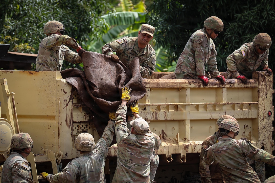 Hawaii National Guardsmen lift debris to fellow guardsmen standing in a hauler with trees in the background.