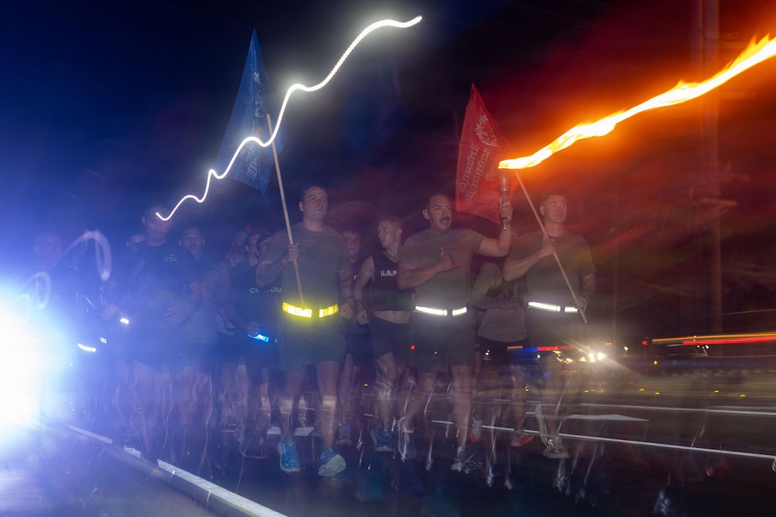 Marines and personnel run in a group in the dark carrying red and blue flags and torches, creating streaks in the sky.