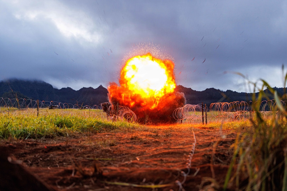 A large explosion in a field near barbed wire creates an orange fireball and clouds of smoke on a gloomy day with mountains in the background.
