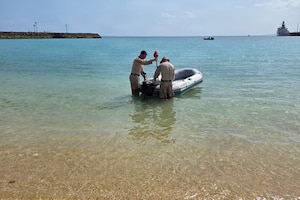 Two men in camouflage military uniforms work on an inflated boat while standing in a shallow body of water.