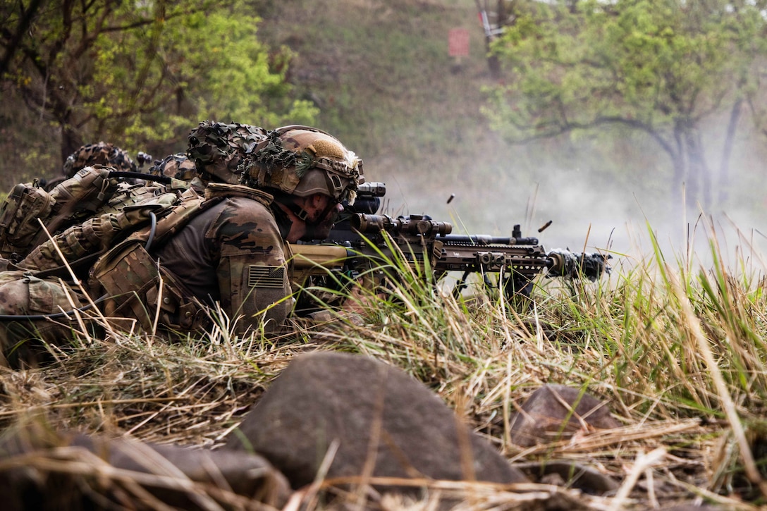 A soldier lying in the grass in a wooded area fires a weapon as ammo flies into the air.