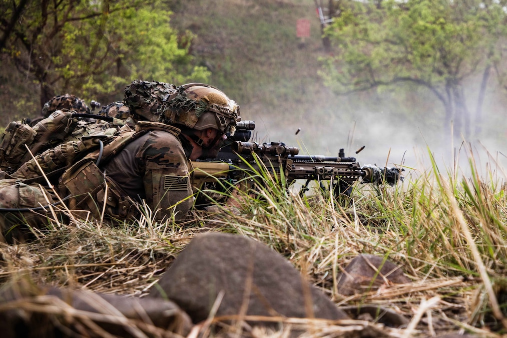 A soldier lying in the grass in a wooded area fires a weapon as ammo flies into the air.