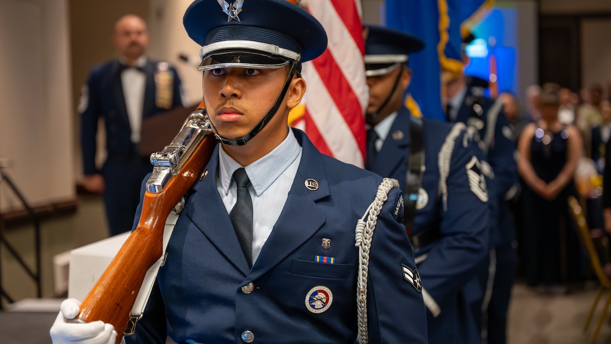 The Airman holding the rifle and leading the Honor Guard is visible in the front left of the frame with the other Airmen out of focus