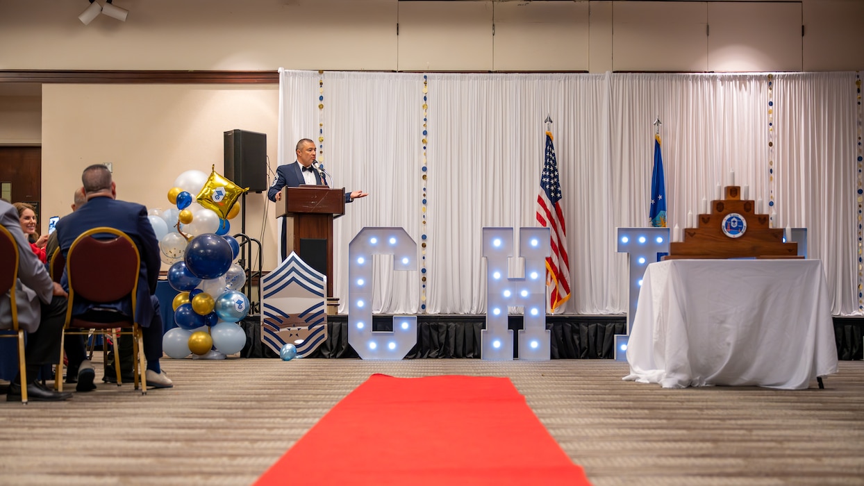 A medium shot of the speaker with audience members and ceremony signage visible