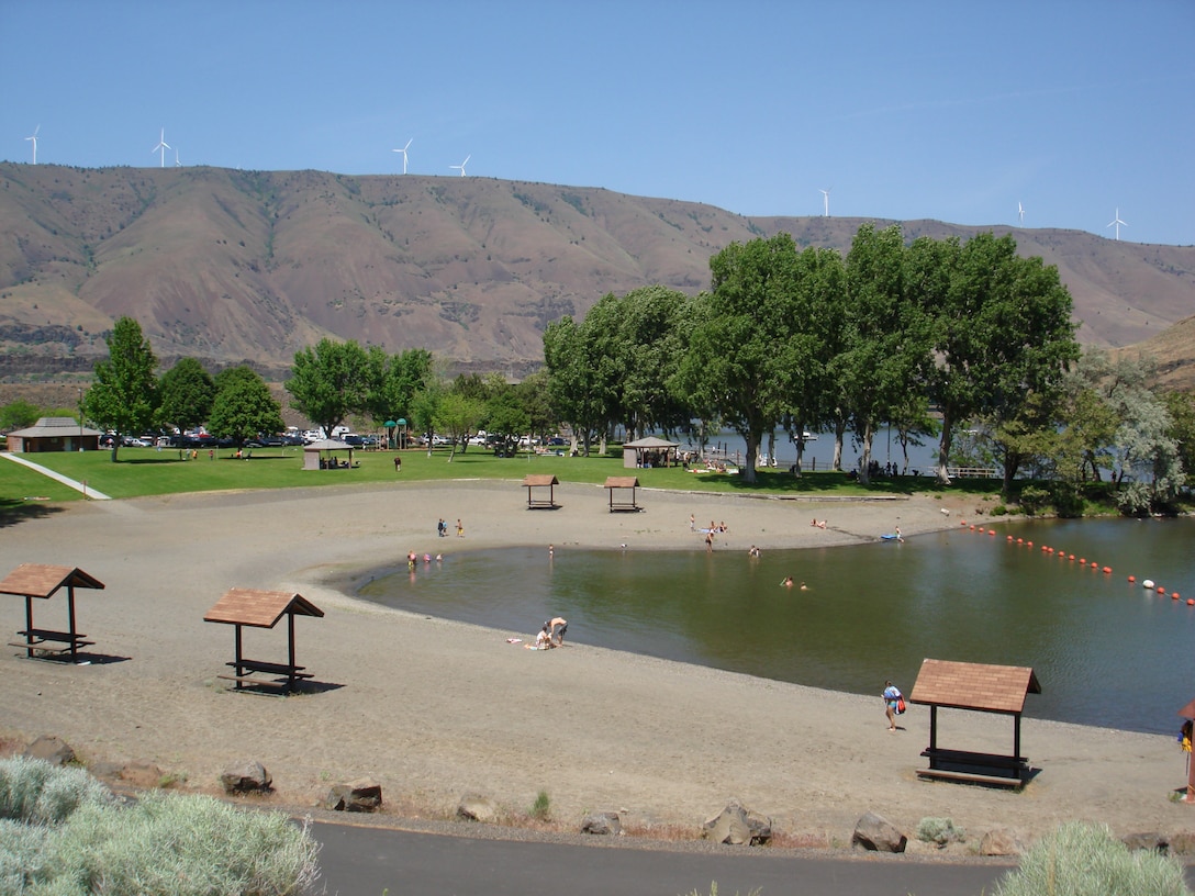 A photos shows a clear blue day – in the distance, near the top of the frame, are large, rugged, steep brown hills that are dotted with green bushes and trees, and topped with large white wind turbines.  In the foreground is a recreation area: a riverside beach, where adults and children are playing. Five covered picnic tables stand empty on the sandy beach area, which is lined by a grassy area that has a bricked building for restrooms and a playground for children. The grassy area has a variety of trees full of green foliage, which gives shade to the visitors who sit below them.