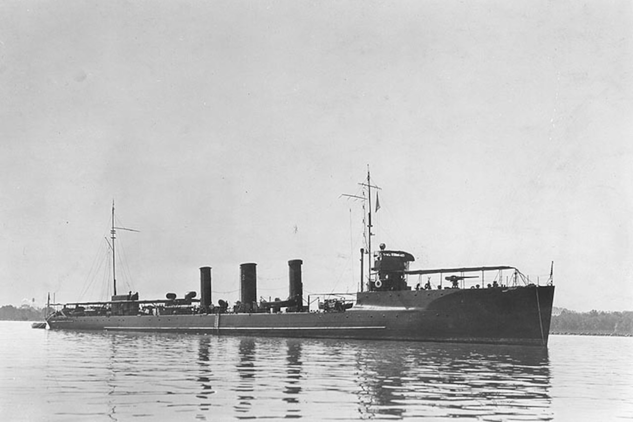 A battleship is anchored in calm water with the shoreline in the distance.