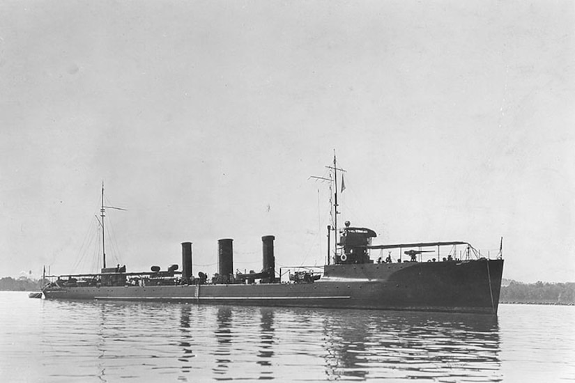 A battleship is anchored in calm water with the shoreline in the distance.