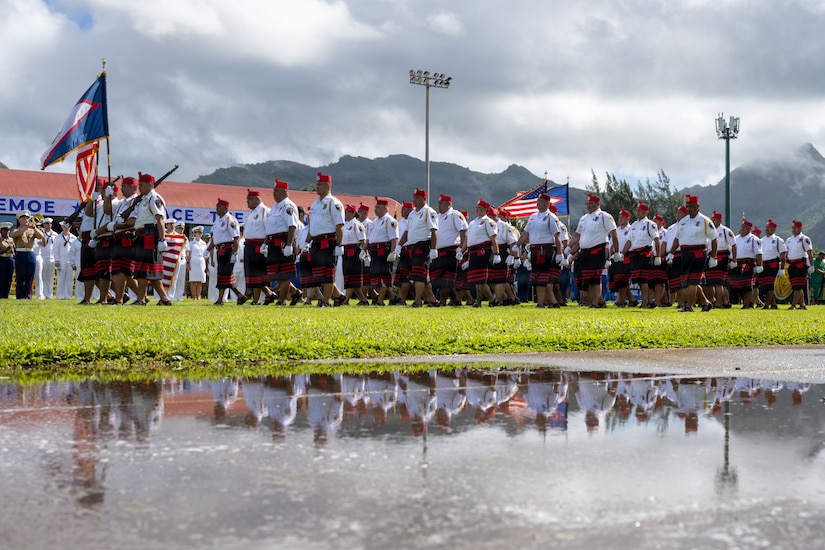 Troops in kilts march on a grassy field with mountains in the background.