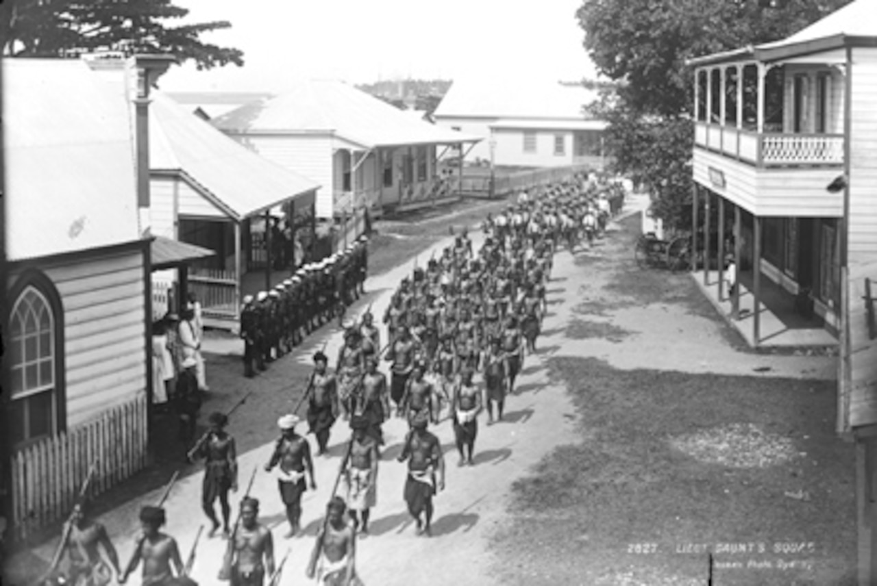 Native warriors with long guns at their right arms march down a dirt road in a village, as a line of soldiers wearing military uniforms stand nearby.