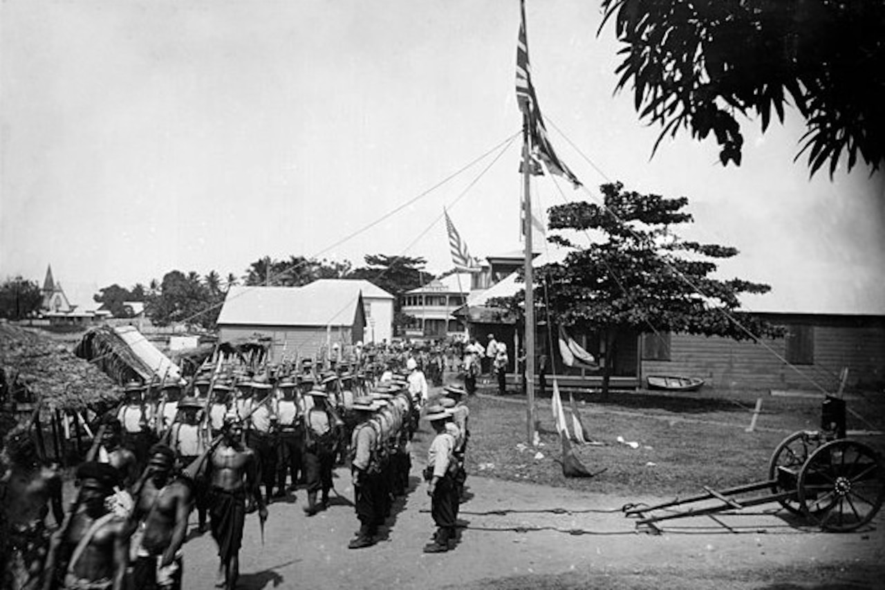 U.S. and British flags fly from a flagpole as troops in uniform and native warriors stand in formation with long guns on a village green.