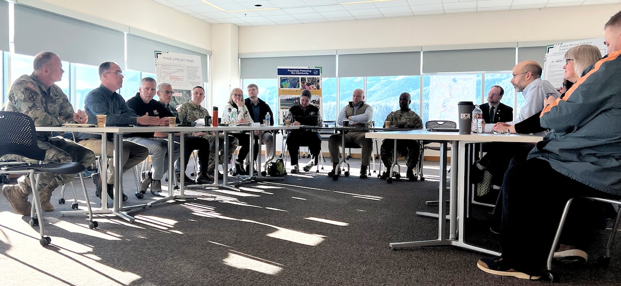 A group of people in civilian attire and camouflage military unforms sit in a meeting room at tables configured in a semicircle.
