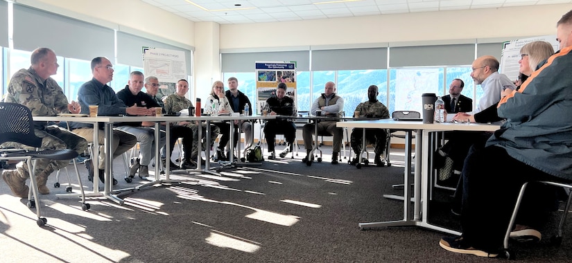 A group of people in civilian attire and camouflage military unforms sit in a meeting room at tables configured in a semicircle.
