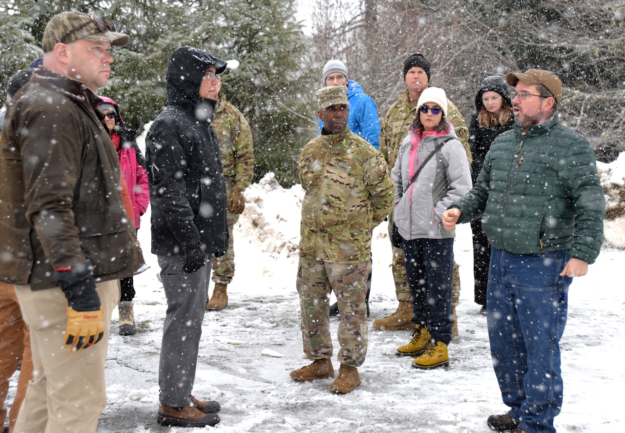 A group of people in civilian attire and camouflage military unforms stand near a wooded area as snow falls listening to a man speak.