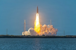 A rocket takes off, with fire and smoke plumes beneath it. The launch pad is adjacent to a wooded area next to a body of water.