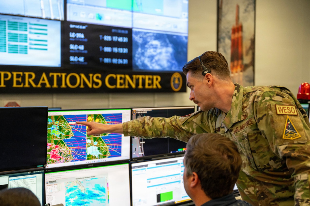 A standing man wearing a camouflage military uniform points to a computer screen. Another man is seated while looking at the same computer monitor.