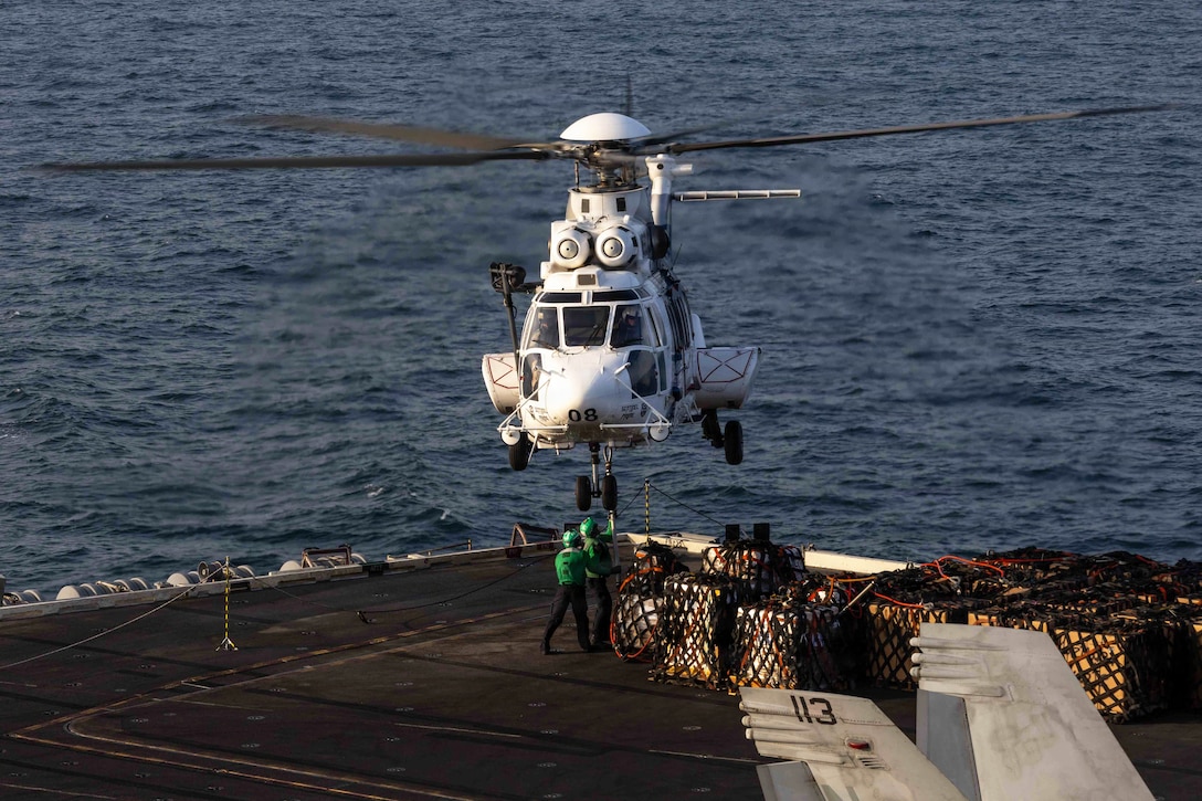 Two sailors in green helmets and vests attach netted cargo from the flight deck of a ship at sea to a hovering helicopter.