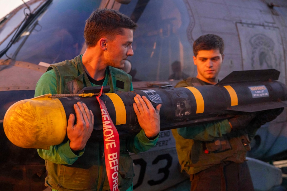 Two sailors in green vests hold a missile in front of a helicopter during the day.