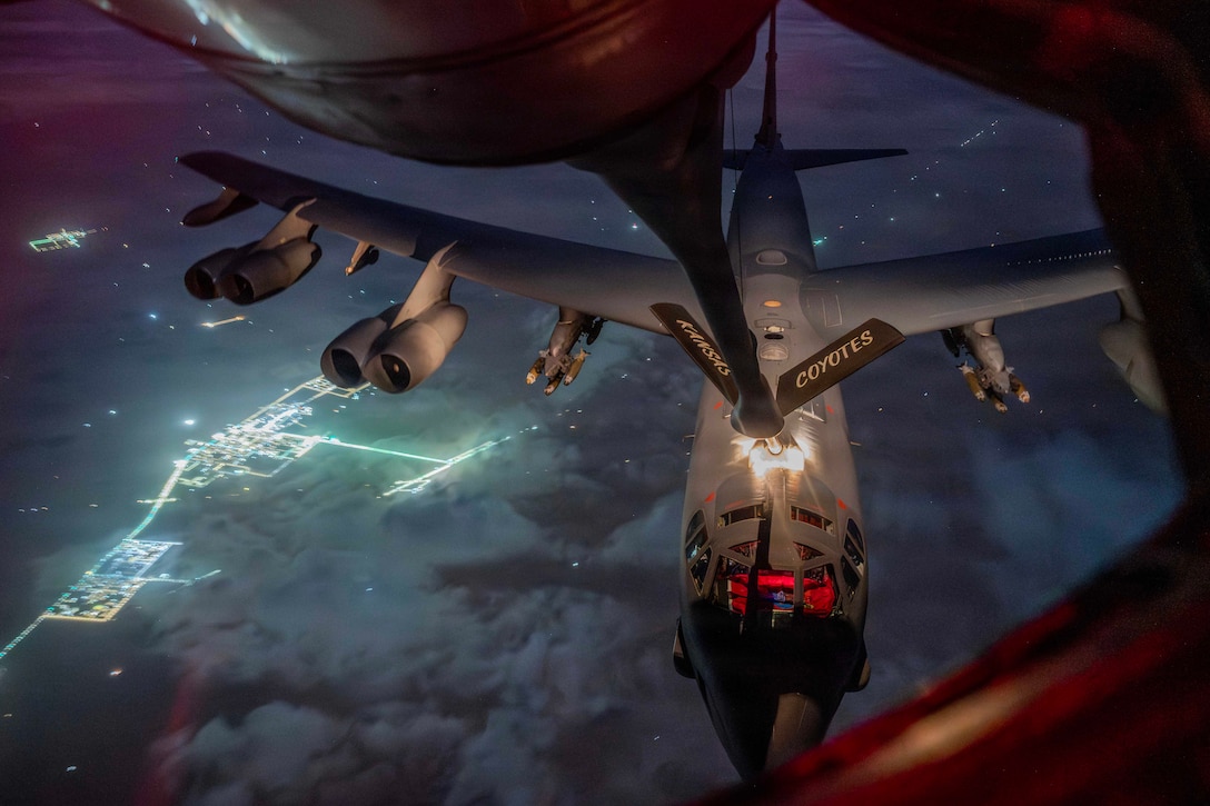 A partially visible aircraft extends its boom to another aircraft in a cloudy, starry night sky over bright city lights.