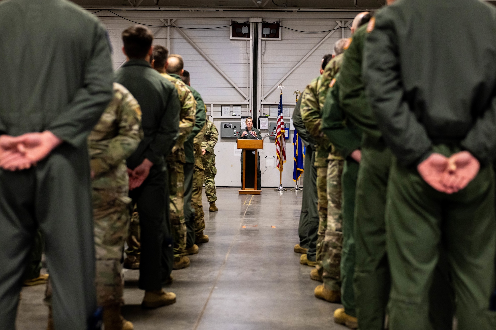 U.S. Air Force Airmen stand in formation while listening to a commander deliver remarks in the front of the room.