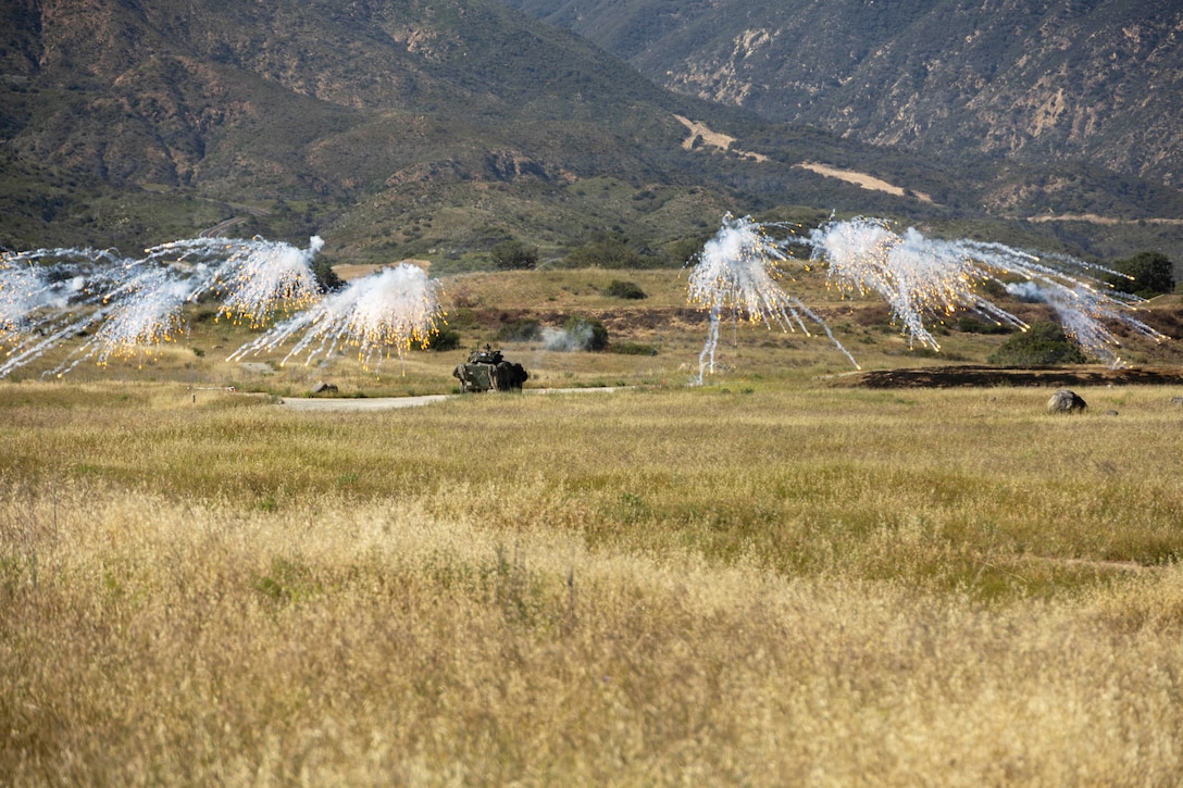 Marines fire a grenade system creating fiery fallen smoke bursts over a field with mountains in the background during the day.