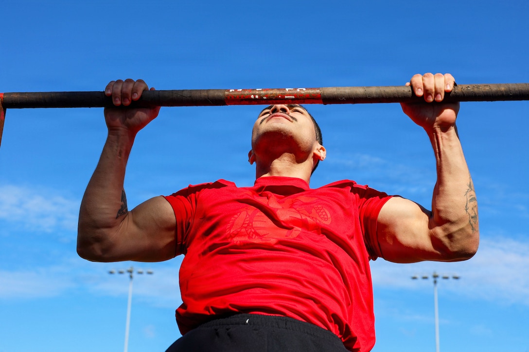A Marine Corps poolee in a red shirt performs a pullup against a blue sky.