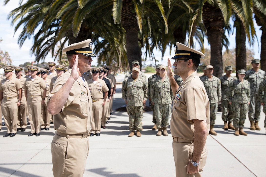 Two sailors face each other with their right hands raised as fellow service members watch while standing in formation under a palm tree during the day.