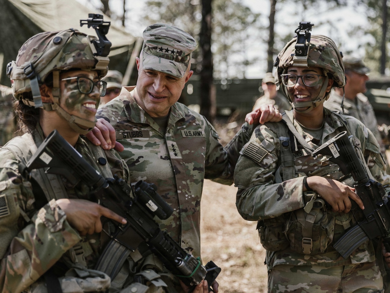 A man wearing a camouflage military uniform talks to two women in similar attire with camouflage face paint who are holding rifles.