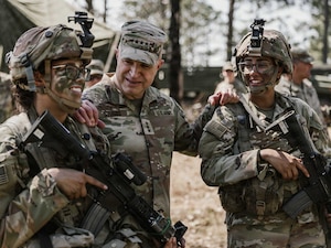 A man wearing a camouflage military uniform talks to two women in similar attire with camouflage face paint who are holding rifles.