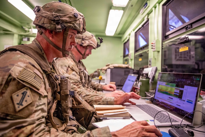 Two men wearing camouflage military uniforms sit in a room working on computers.