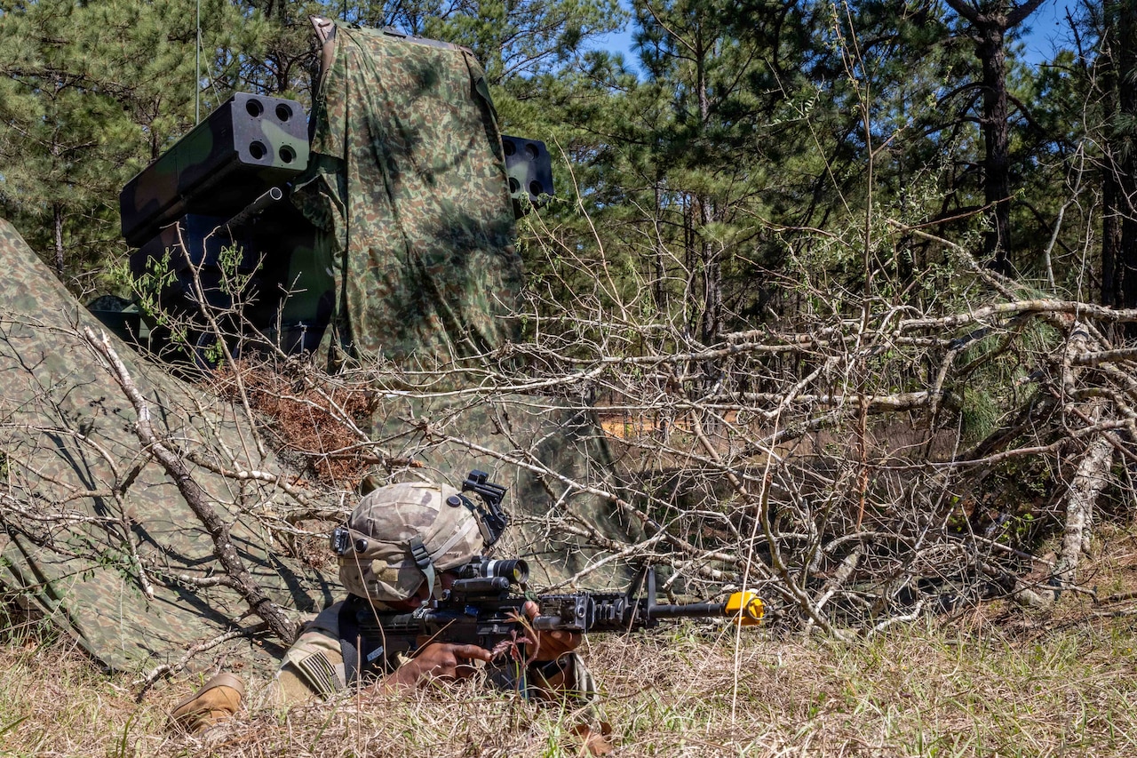 A person wearing a camouflage military uniform lies on the ground in the woods while aiming a rifle. There is a military air defense system with a camouflage sheet draped over it in the background.