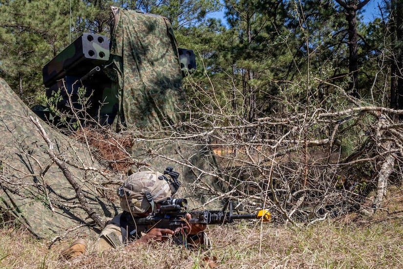 A person wearing a camouflage military uniform lies on the ground in the woods while aiming a rifle. There is a military air defense system with a camouflage sheet draped over it in the background.