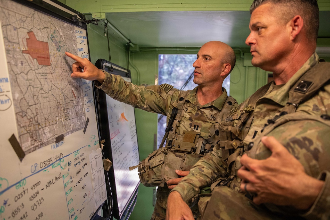 A man wearing a camouflage military uniform stands in a room pointing at a location on a map hanging from a whiteboard while another man in similar attire looks at the map.
