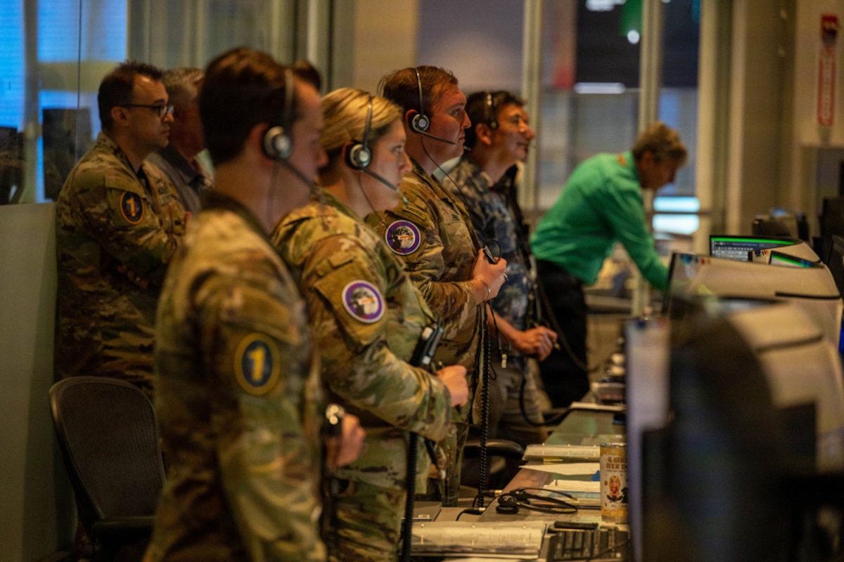 Several people wearing camouflage military uniforms and headsets stand in front of computer screens. A man in the background is dressed in civilian clothing.