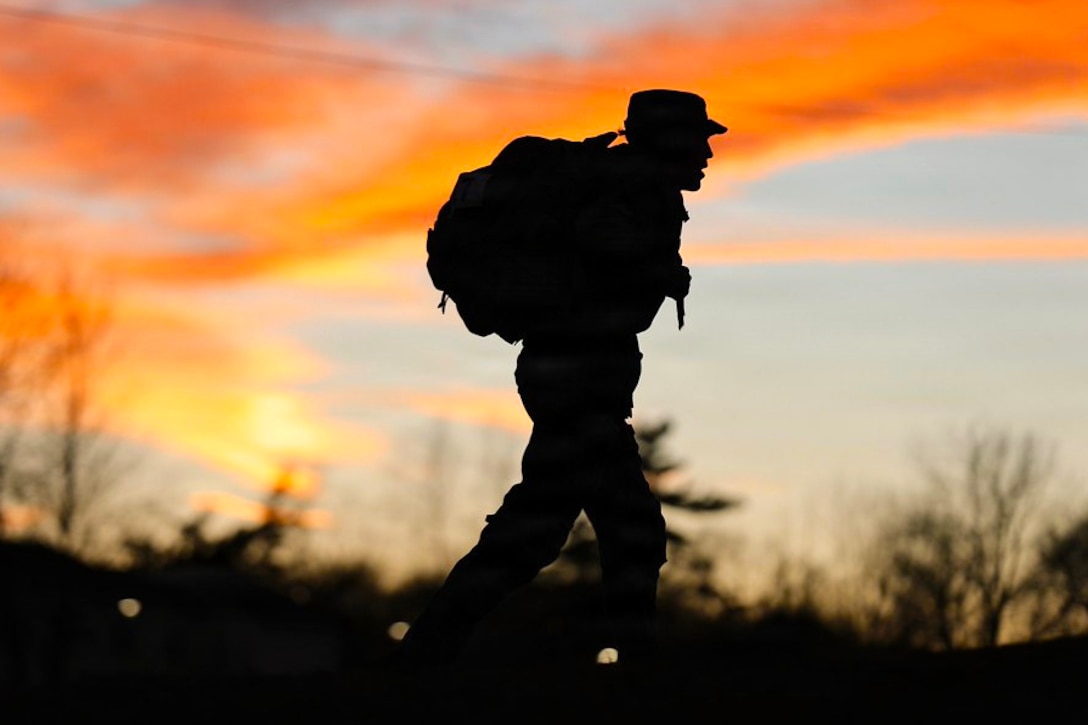 A soldier in silhouette carrying a rucksack walks outside, with the sun and trees without leaves in the background.