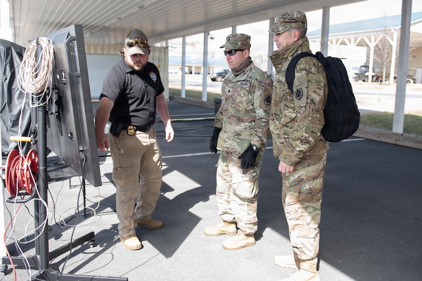 A man wearing civilian attire speaks as he points to a board while two men dressed in camouflage military uniforms look at the board.