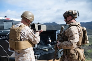 Two service members wearing camouflage military uniforms, helmets and sunglasses stand at the bed of a pickup truck while the man on the left is holding a piece of military equipment.