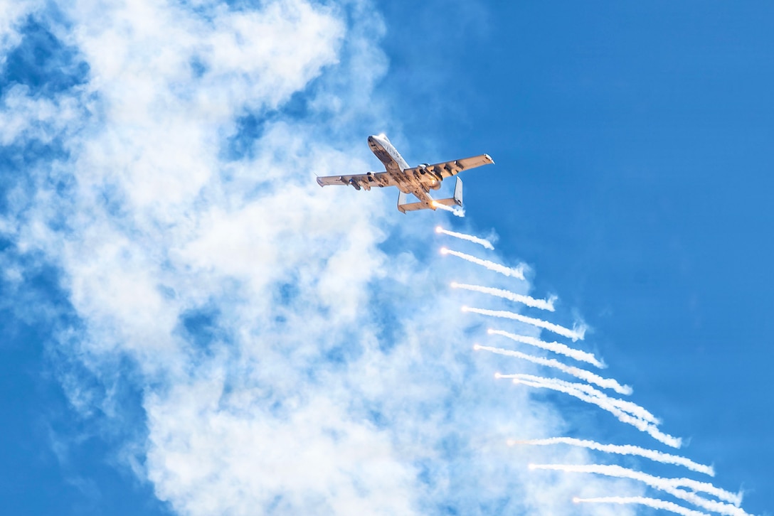 An aircraft deploying flares is seen against a blue sky and bright white clouds.