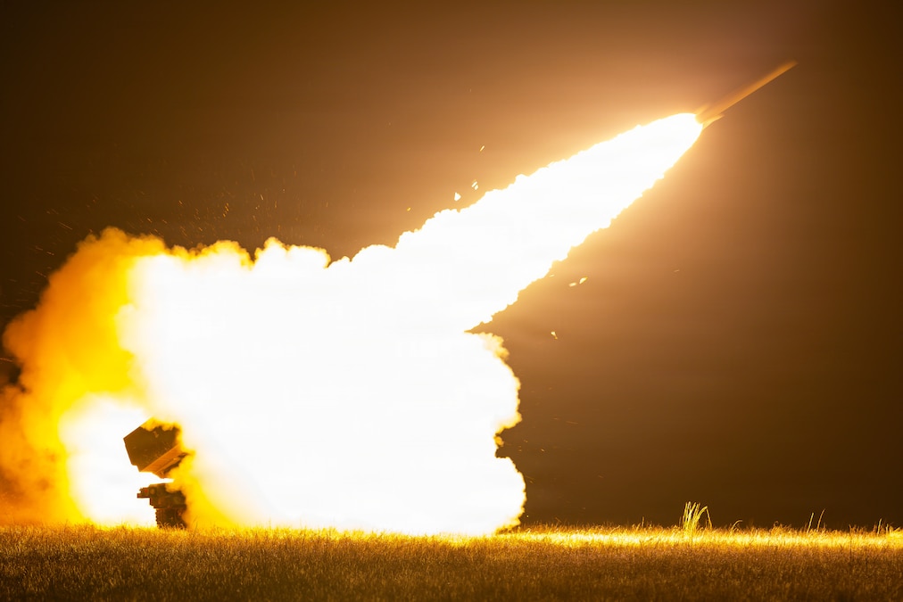 A rocket is airborne above a field at night, with a trail of fire and smoke behind it.