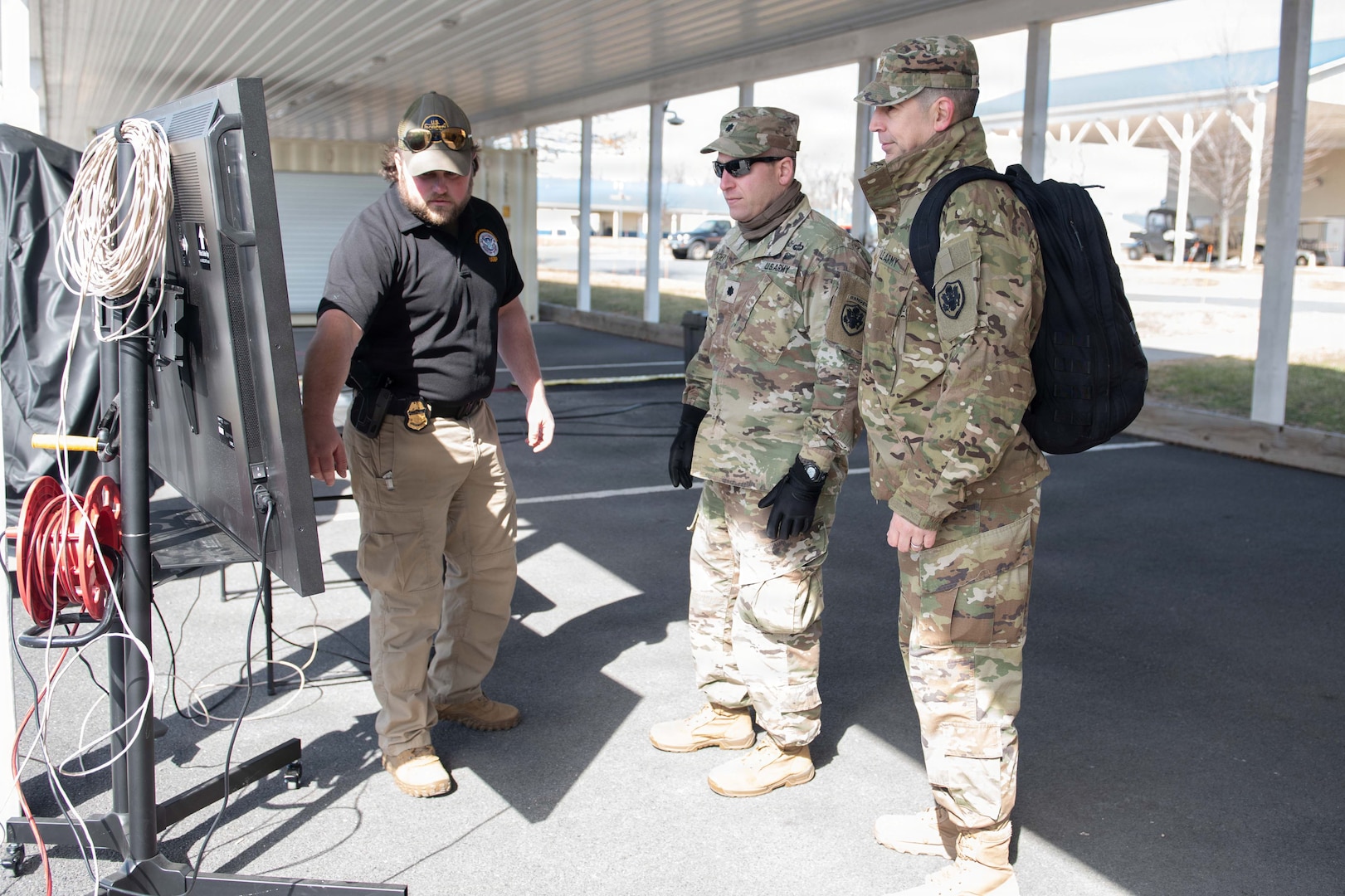 A man wearing civilian attire speaks as he points to a board while two men dressed in camouflage military uniforms look at the board.