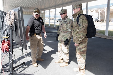 A man wearing civilian attire speaks as he points to a board while two men dressed in camouflage military uniforms look at the board.
