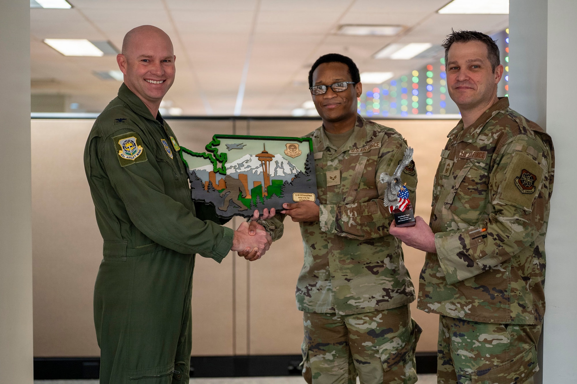 three U.S. Air Force airman face the camera in smile, the one on the left is in a flight, while the other two are in camouflage. The airman on the left is handing a plaque to the airman in the center