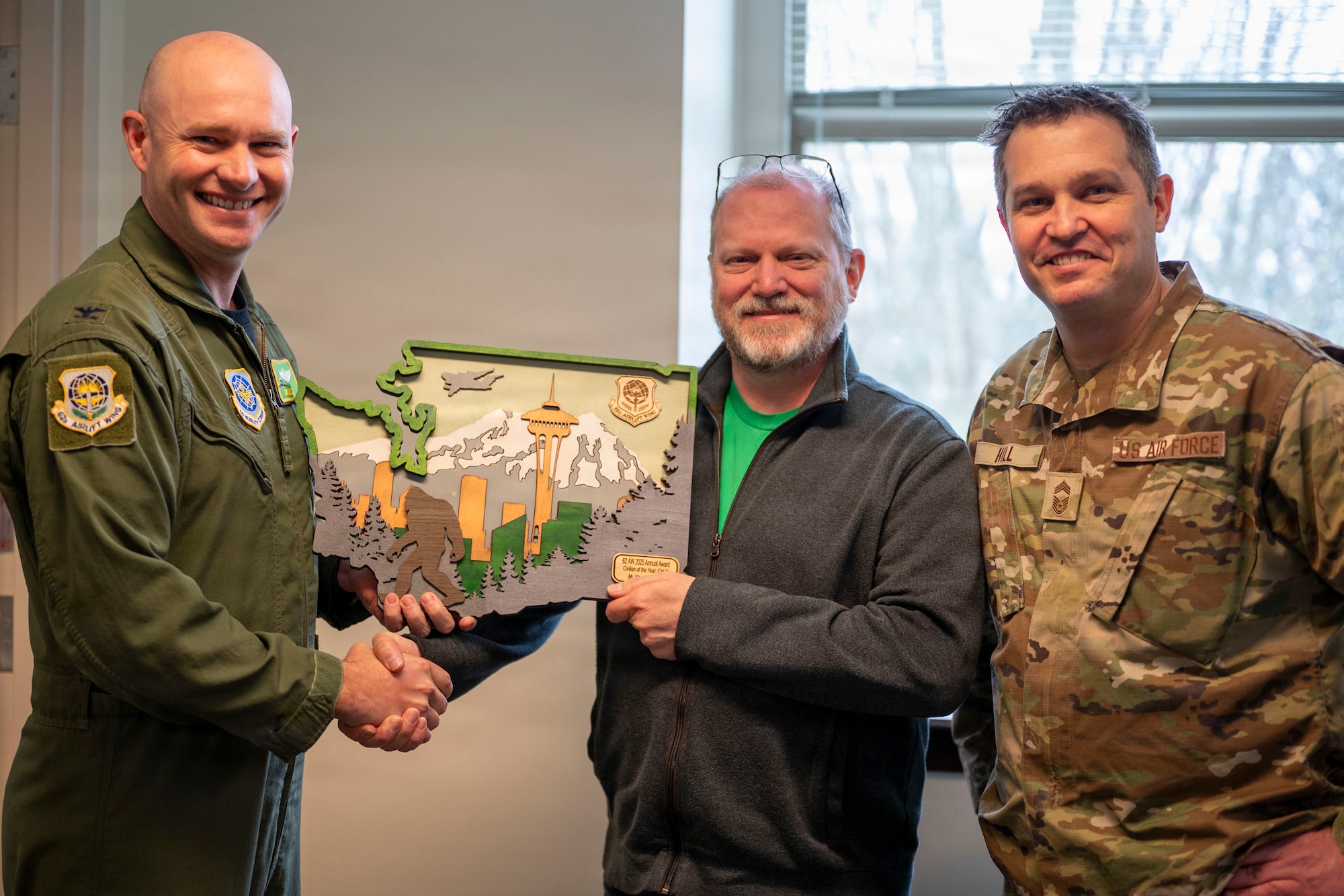 two U.S. Air Force airman stand on either side of a male civilian, all facing the camera and smiling. The airman on the left hands a plaque to the airman in the center